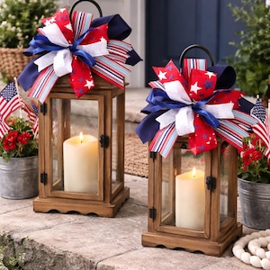 May include: Two wooden lanterns with glass panels, each holding a lit candle. The lanterns are decorated with red, white, and blue bows. Small American flags and potted flowers are in the background, creating a patriotic theme.