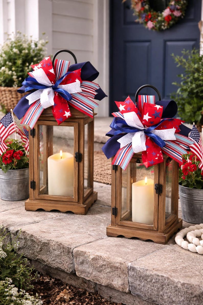 May include: Two wooden lanterns with glass panels, each holding a lit candle. The lanterns are decorated with red, white, and blue bows. Small American flags and potted flowers are in the background, creating a patriotic theme.