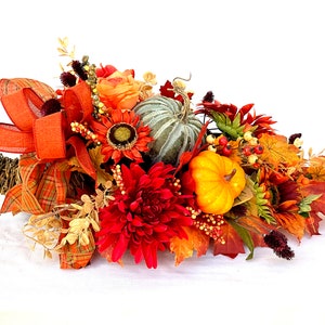 May include: A fall-themed cornucopia arrangement featuring a variety of artificial flowers, pumpkins, and leaves in shades of orange, red, and yellow. A large orange plaid ribbon adds a decorative touch to the arrangement.