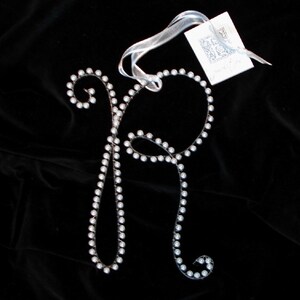 May include: A silver letter R made of wire and decorated with white pearls. The letter is hanging from a white ribbon with a small white tag that says "My initials".