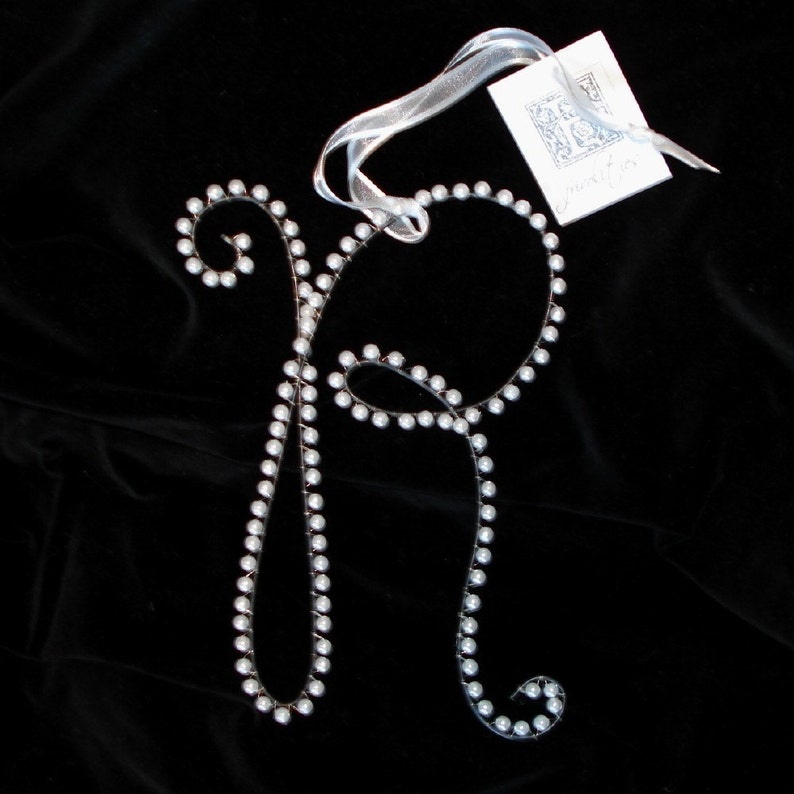 May include: A silver letter R made of wire and decorated with white pearls. The letter is hanging from a white ribbon with a small white tag that says "My initials".