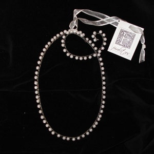 May include: A white pearl necklace with a simple oval shape. The necklace is on a black background with a white ribbon and a tag that says "frivolites".