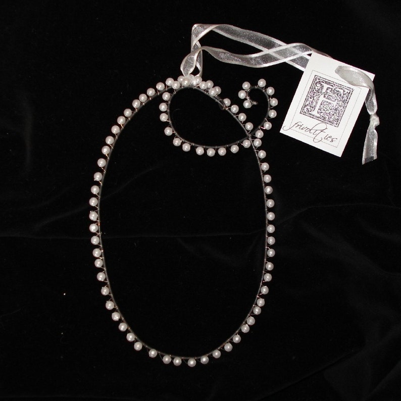 May include: A white pearl necklace with a simple oval shape. The necklace is on a black background with a white ribbon and a tag that says "frivolites".