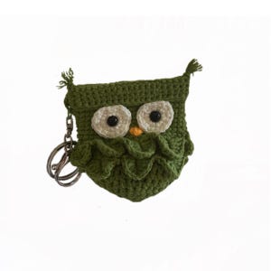 May include: A handmade green crochet owl coin purse with a key ring. The owl has large, off-white eyes with black pupils and a small orange beak. The purse is made of a textured yarn and has a flap closure.