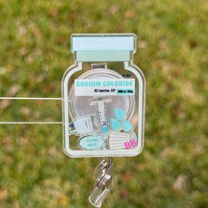 May include: A clear, rectangular badge reel with a medical theme. The design features a vial labeled "SODIUM CHLORIDE" with a syringe, medical mask, and other medical elements. The reel has a retractable cord and a metal clip.