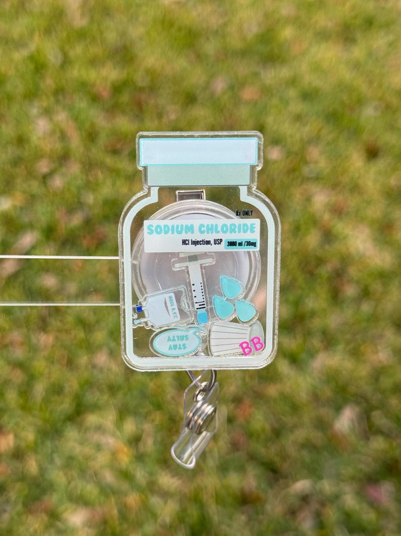 May include: A clear, rectangular badge reel with a medical theme. The design features a vial labeled "SODIUM CHLORIDE" with a syringe, medical mask, and other medical elements. The reel has a retractable cord and a metal clip.