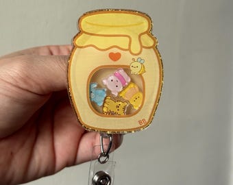 Puede incluir: Un portatarjetas con forma de tarro de miel amarillo con un borde dorado brillante. Dentro del tarro hay personajes de dibujos animados, como un cerdo rosa, un oso azul, un tigre amarillo y una abeja. El portatarjetas tiene una correa de plástico transparente y un clip de metal.