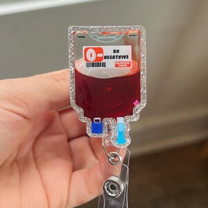 May include: A clear, rectangular badge reel with a glittery border, featuring a blood bag design. The bag is filled with red liquid and has the text "O- RH NEGATIVE VOLUNTEER DONOR". The reel has a clear plastic clip and a retractable cord.