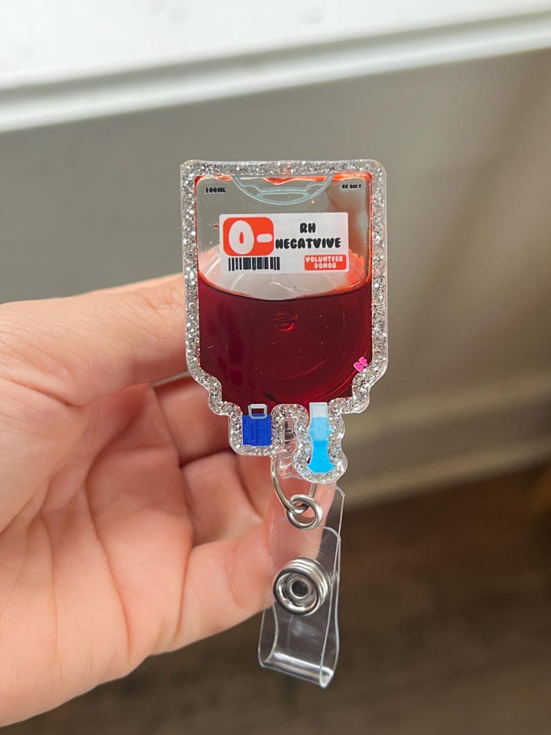 May include: A clear, rectangular badge reel with a glittery border, featuring a blood bag design. The bag is filled with red liquid and has the text "O- RH NEGATIVE VOLUNTEER DONOR". The reel has a clear plastic clip and a retractable cord.