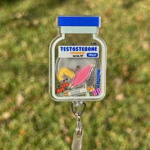 May include: A clear, acrylic badge reel featuring a cartoon vial design with the word "TESTOSTERONE" in blue and white. The reel includes a syringe, muscle graphic, and the text "200mg/ml". The reel has a blue top and a silver clip.
