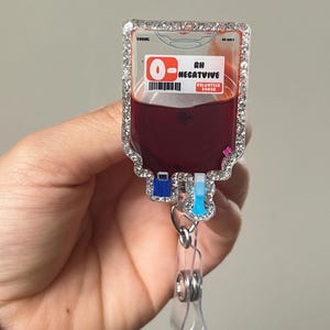 May include: A badge reel shaped like a blood bag, with a clear, sparkly border. The bag is filled with a dark red liquid and has the text "O- Negative Volunteer Donor". The reel has a clear plastic clip.