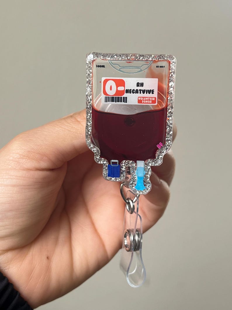May include: A badge reel shaped like a blood bag, with a clear, sparkly border. The bag is filled with a dark red liquid and has the text "O- Negative Volunteer Donor". The reel has a clear plastic clip.