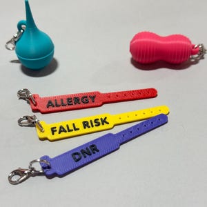 May include: A collection of five 3D printed charms including a blue bulb syringe, a pink peanut shape, and three medical identification wristband replicas in red, yellow, and purple. The wristbands feature the black text ALLERGY, FALL RISK, and DNR.