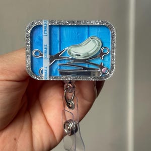 May include: A rectangular badge reel with a glittery silver border. The badge reel features a blue background with a medical instrument design. The design includes a kidney-shaped object, scissors, and tweezers. The text "STERILE-Z" is printed on the side.