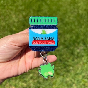 Peut inclure: Un porte-badge bleu pailleté avec les mots "SANA SANA COLITA DE RANA" en blanc et rouge. Le porte-badge a un motif de grenouille verte et un charm de grenouille transparent attaché.