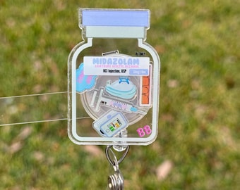 May include: A clear acrylic badge reel in the shape of a medicine vial, featuring the text "MIDAZOLAM" and medical illustrations. The badge reel has a retractable cord and a metal clip for attaching to clothing.
