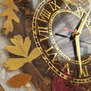 May include: A brown and gold clock face with Roman numerals and gold hands. The clock face is surrounded by fall leaves in shades of gold, red, and brown. The clock is encased in clear resin.