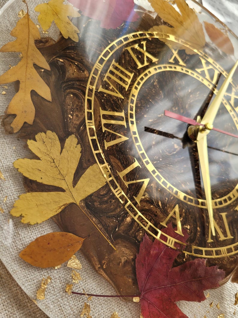 May include: A brown and gold clock face with Roman numerals and gold hands. The clock face is surrounded by fall leaves in shades of gold, red, and brown. The clock is encased in clear resin.