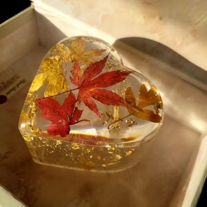 Op de afbeelding: Een hartvormige harsdoos met ingebedde herfstbladeren in tinten rood en goud, en goudvlokken. De heldere hars maakt de bladeren zichtbaar. De doos zit in een lichtgekleurde doos.