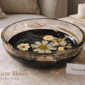 May include: A decorative glass bowl with a black interior, featuring dried flowers and gold leaf accents. The bowl is part of the "Nocturne Bloom Collection" and has a clear rim. A small white card reads "Nocturne Bloom I".