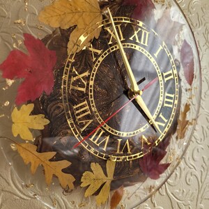 May include: A round, clear resin clock with gold Roman numeral markings and gold hands. The clock face is embedded with dried leaves in shades of red, yellow, and brown.