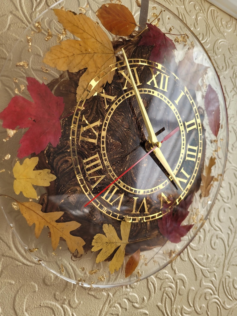 May include: A round, clear resin clock with gold Roman numeral markings and gold hands. The clock face is embedded with dried leaves in shades of red, yellow, and brown.