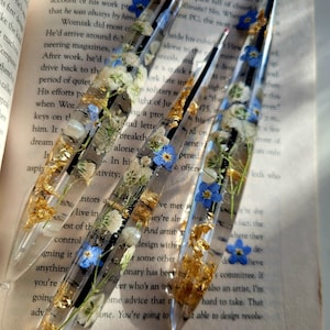May include: Three clear resin pens featuring embedded dried blue forget-me-not flowers, small white baby's breath, and gold leaf flakes. The pens have a tapered shape with a pointed tip and are displayed resting on an open book page with text.