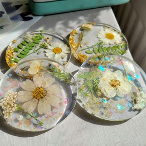 Puede incluir: Cuatro posavasos redondos de resina transparente con flores secas y pan de oro. Cada posavasos presenta un arreglo floral diferente, incluyendo margaritas y otras flores delicadas, con detalles de purpurina iridiscente.