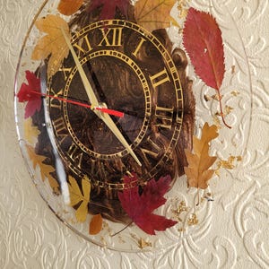 May include: A round clock with Roman numerals and gold accents. The clock face is brown with a marbled effect. The clock is encased in clear resin with dried leaves and gold flakes.