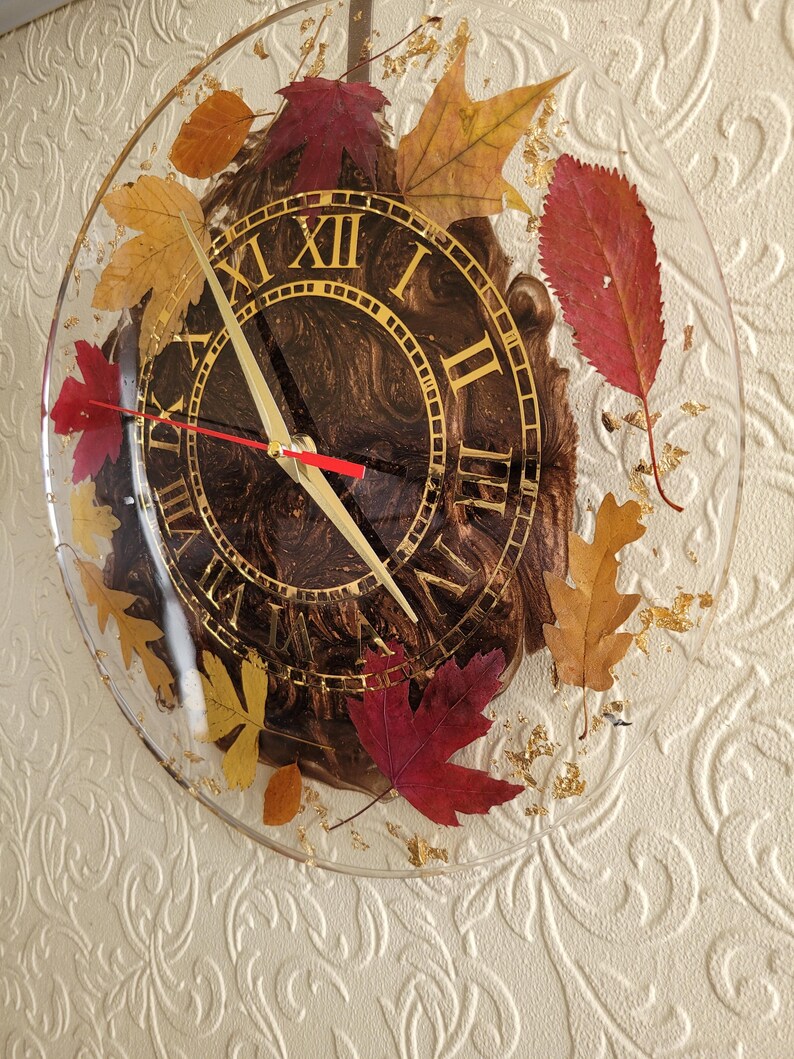 May include: A round clock with Roman numerals and gold accents. The clock face is brown with a marbled effect. The clock is encased in clear resin with dried leaves and gold flakes.