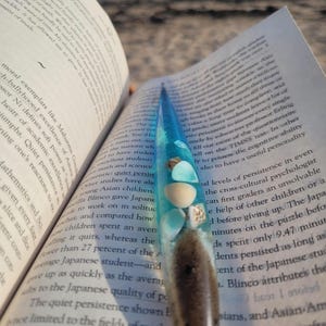 Op de afbeelding: Een handgemaakt boekenlegger met een harsontwerp. De boekenlegger is transparant met blauwe en bruine tinten en bevat kleine schelpen en zand. Het ligt in een open boek met zichtbare tekst.