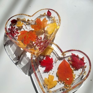 Puede incluir: Dos bandejas de resina en forma de corazón. Cada bandeja es transparente con hojas de otoño incrustadas en tonos rojos, naranjas y amarillos, con copos de oro. Las bandejas están sobre una superficie blanca, proyectando sombras.