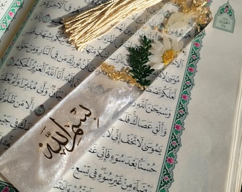May include: A clear resin bookmark featuring embedded white daisy flowers, green foliage, and gold flakes. The bookmark has a gold tassel at the top and the Arabic calligraphy "Bismillah" written in black ink on the lower portion of the surface.