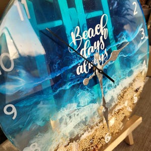 Puede incluir: Un reloj redondo con una escena de océano azul y blanco. La esfera del reloj tiene números blancos y las palabras "Beach days always" están escritas en blanco en la esfera del reloj. El reloj está en un caballete de madera.