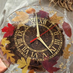 May include: A round clock with Roman numerals and gold accents. The clock face is made of brown resin with gold flecks and features real pressed leaves.
