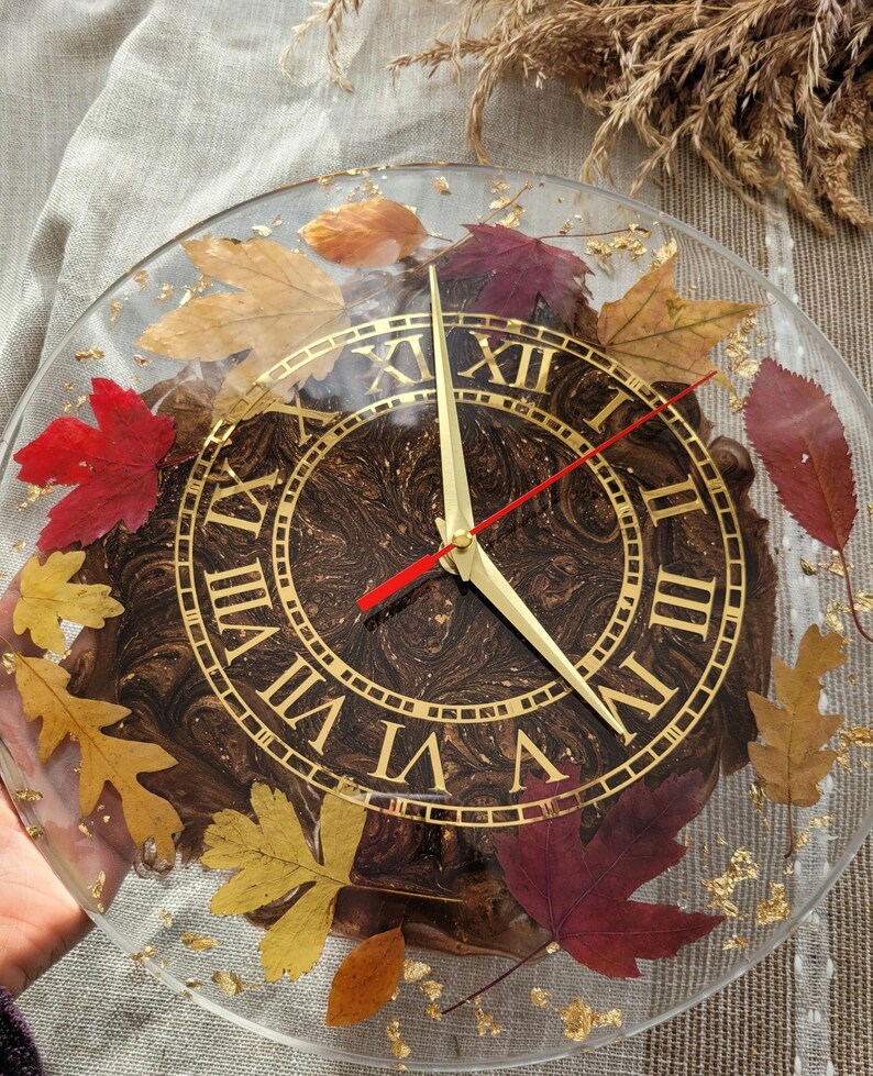 May include: A round clock with Roman numerals and gold accents. The clock face is made of brown resin with gold flecks and features real pressed leaves.