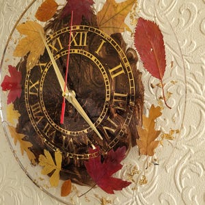 Puede incluir: Un reloj redondo con números romanos y detalles dorados. La esfera del reloj es marrón con un efecto marmoleado. El reloj está rodeado de hojas secas en tonos rojos, naranjas y amarillos.