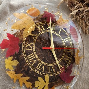 May include: A round, clear resin clock face with Roman numeral markings and gold accents. The clock face is embedded with dried leaves in shades of red, orange, and yellow.