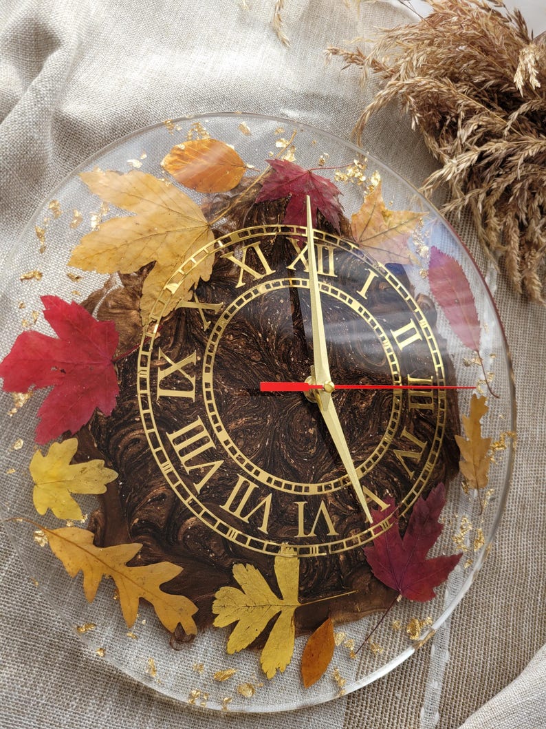 May include: A round, clear resin clock face with Roman numeral markings and gold accents. The clock face is embedded with dried leaves in shades of red, orange, and yellow.