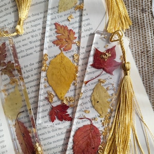 May include: Four bookmarks with gold tassels and pressed autumn leaves encased in clear resin. The leaves are shades of red, orange, and yellow with gold leaf accents.