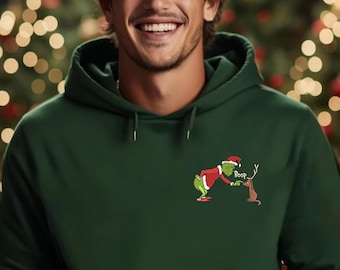 May include: A dark green hoodie featuring a Christmas-themed graphic of a green character in a Santa hat and a reindeer. The hoodie has a drawstring hood and a kangaroo pocket. The background includes blurred Christmas trees with lights.