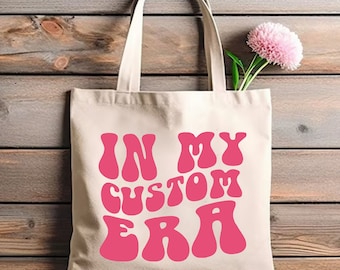 May include: A white canvas tote bag with pink text that reads "In My Custom Era". The text is in a retro, groovy font style.