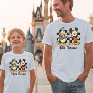 May include: White t-shirts with a cartoon graphic of Donald Duck and Mickey Mouse on a checkered background. The shirt on the left says "Little Trouble" and the shirt on the right says "Big Trouble".