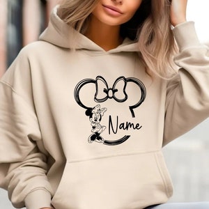 K&ouml;nnte beinhalten: Ein beiger Hoodie mit einem schwarz-wei&szlig;en Aufdruck von Minnie Mauss Kopf mit Schleife und dem Wort "Name" darunter.