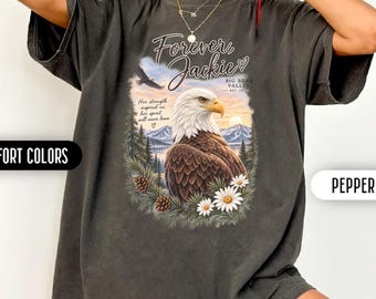 May include: A pepper-colored Comfort Colors t-shirt featuring a graphic of a bald eagle, pine trees, and mountains. The text reads Forever Jackie, Big Bear Valley, Est 2024, and Her strength inspired us, her spirit will never leave.