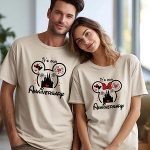 May include: Two beige t-shirts featuring a black Mickey Mouse head design. The design includes a castle, hearts, and the text "It's our 1st Anniversary". One shirt has a red bow, the other has a red heart.