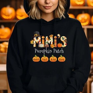 May include: A black hooded sweatshirt featuring a graphic print that reads Mimi's Pumpkin Patch. The design includes a pilgrim hat, autumn leaves, and four orange pumpkins with the names Charlotte, Riley, St. Pat, and Alivia printed on them.