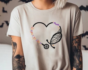 May include: A light beige short-sleeved t-shirt featuring a graphic of a black heart outline combined with a stethoscope. The design includes a spiderweb pattern, a small purple bat, and the text "SPOOKY SEASON" in colorful letters.