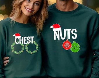 May include: Two dark green crewneck sweatshirts featuring holiday-themed graphics. One sweatshirt displays the word CHEST in white with two green wreaths. The other sweatshirt displays the word NUTS in white with a red and a green ornament.
