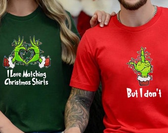 May include: Two matching Christmas shirts. One green shirt with a Grinch hand making a heart shape with Christmas lights and the text "I love matching Christmas shirts". The other red shirt has a Grinch hand making a middle finger gesture with Christmas lights and the text "But I don't".